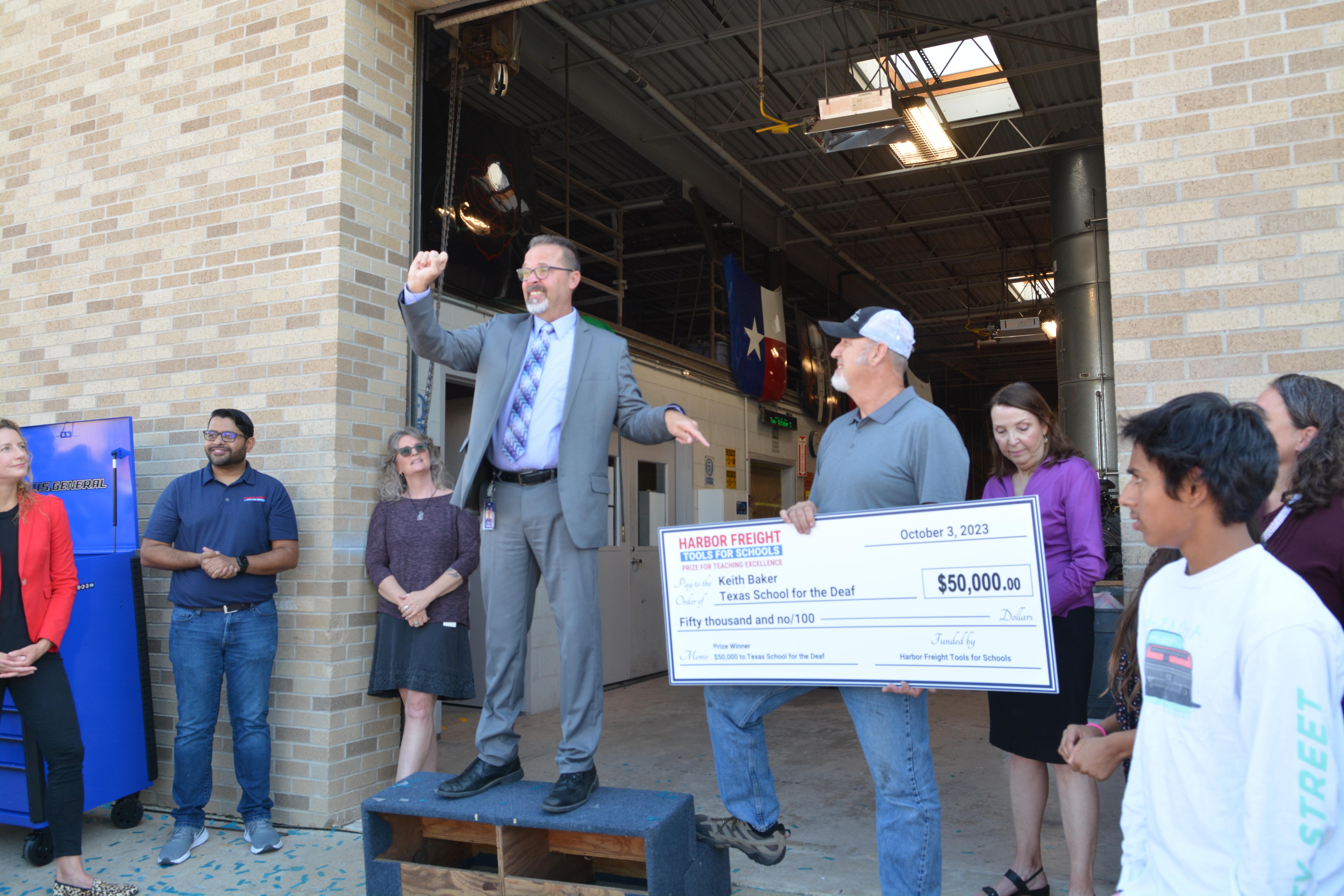 People are standing behind the warehouse, Peter Bailey welcomes Keith Baker with a big check.
