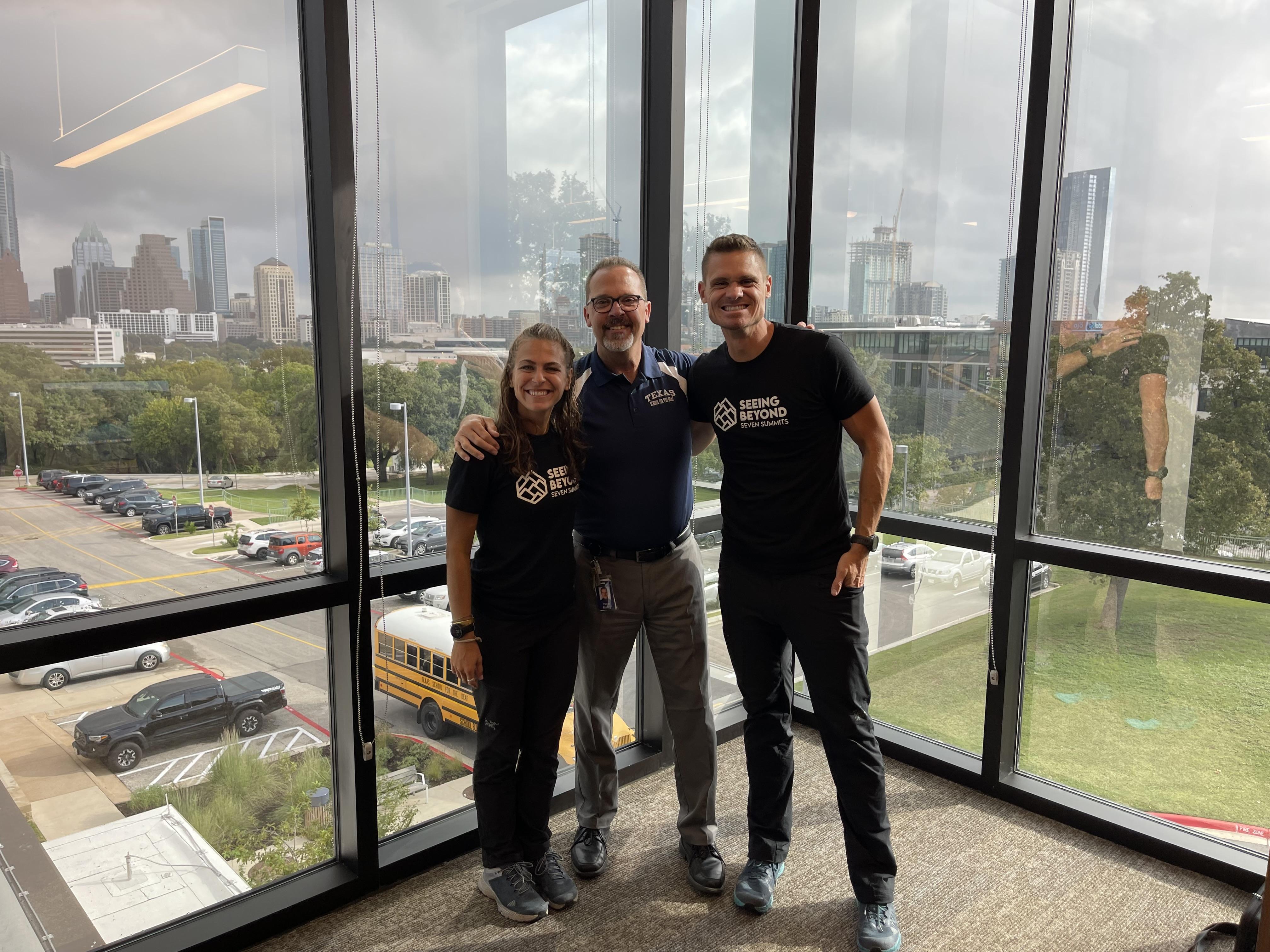 Peter Bailey is standing between two deaf mountaineers, wearing TSD blue polo shirt and gray pants. The deaf mountaineers wore black shirts with their logo on the middle and the text: Seeing Beyond Seven Summits. The background is Austin's skyline with cloudy weather.