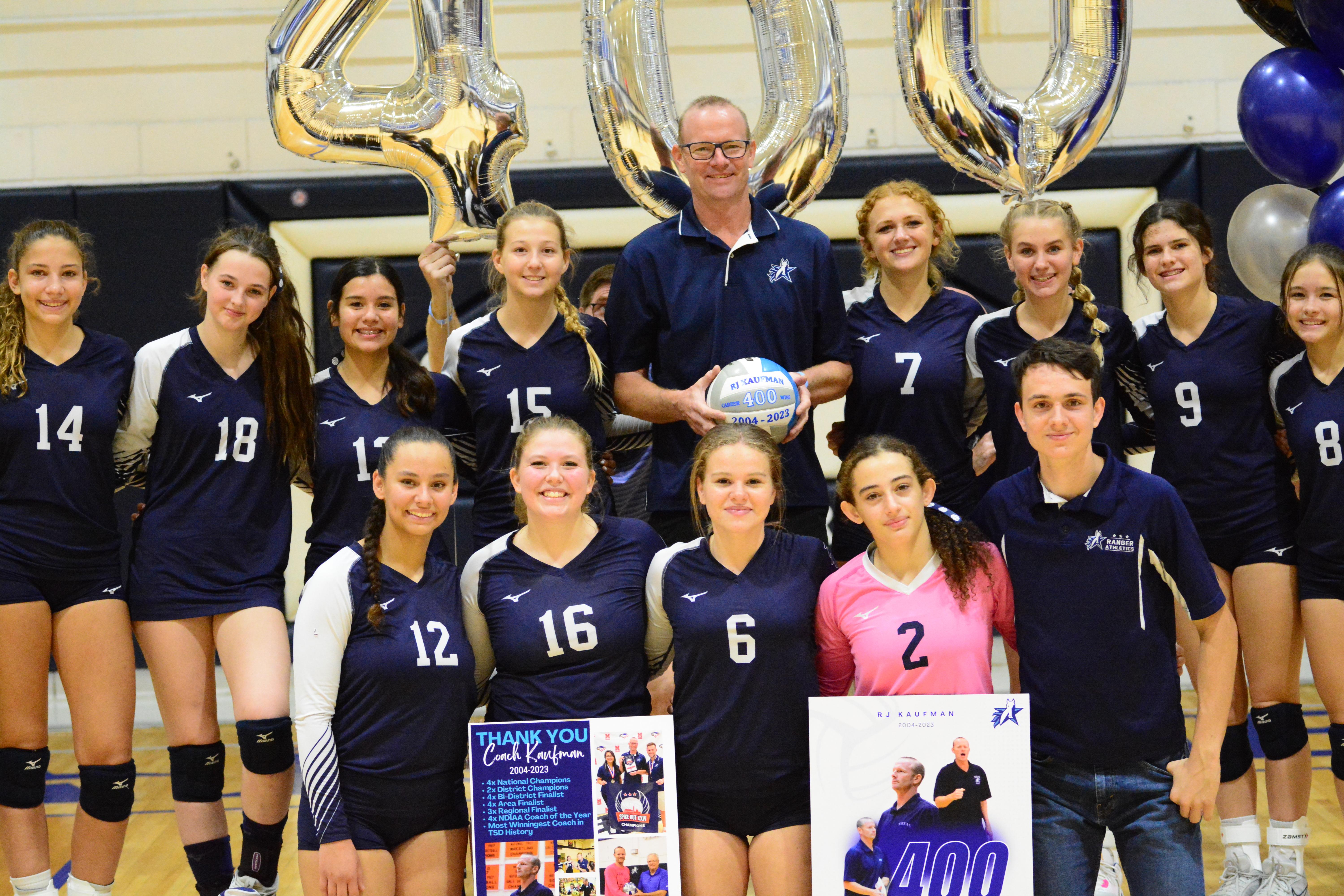 Coach RJ Kaufman posed in middle with his volleyball athletes, smiling. He wore coach navy blue polo shirt and holding the volleyball and the text reads: RJ Kaufman career 400 wins 2004-2023. All the volleyball athletes wore the uniforms with numbers on the front. The background has 3 silver balloons that reads "400." The front volleyball players held two posters of RJ Kaufman. 