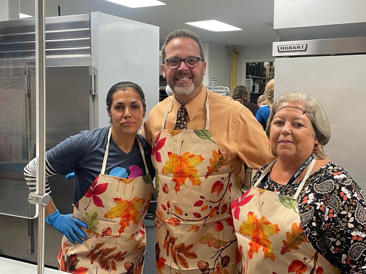 Peter Bailey posed with two cafeteria staff, all of them are wearing fall leaves aprons and smiling. 