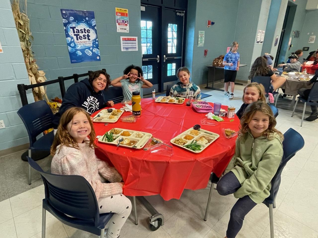 The five elementary students sat around the cafeteria table and smiling towards the camera. A staff sat with them as well, smiling to the camera. They all have delicious lunch trays front of them.