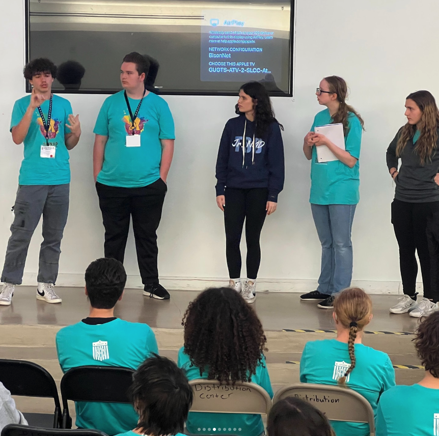 A student talked on the stage during the workshop, other 4 students looking at the student who talked. 3 people wore bright blue shirts, one person wore Jr. NAD sweater, and another person wore gray long sleeves jacket. 