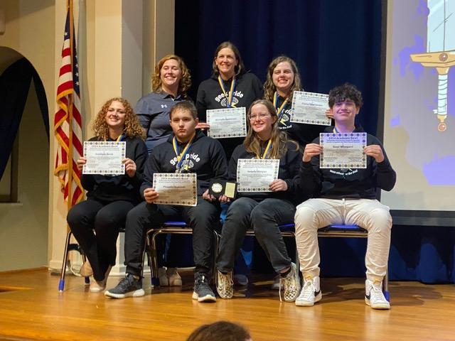 4 students sitting on the front, posed with award along with three adults behind the students. They all wear dark hoodies.