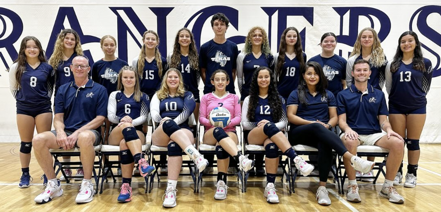 The High School Varsity volleyball team posed for the team picture. 11 people stand in the back, 7 people sit in the front. They are wearing long sleeved blue and white uniforms with numbers on the front. The head coach is sitting on the far left, the two remaining assistant coaches are sitting on the far right.