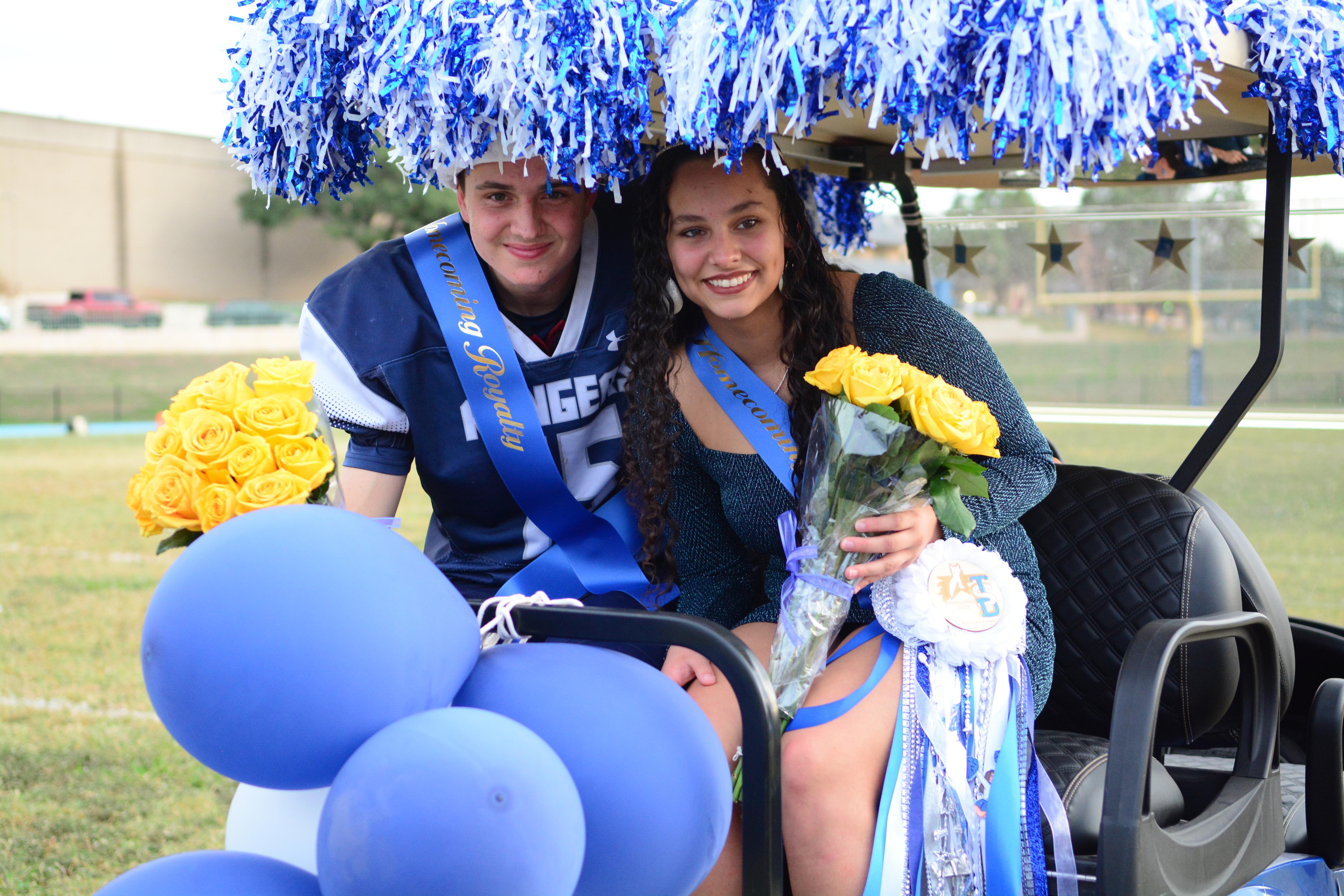 Two Homecoming Royalty, Xavier and McKenna, posed sitting in the back of the golf cart decorated with blue and white streamers and balloons. Both of them have stash’s text: Homecoming Royalty. They held their bouquet of yellow flowers and they smiled.