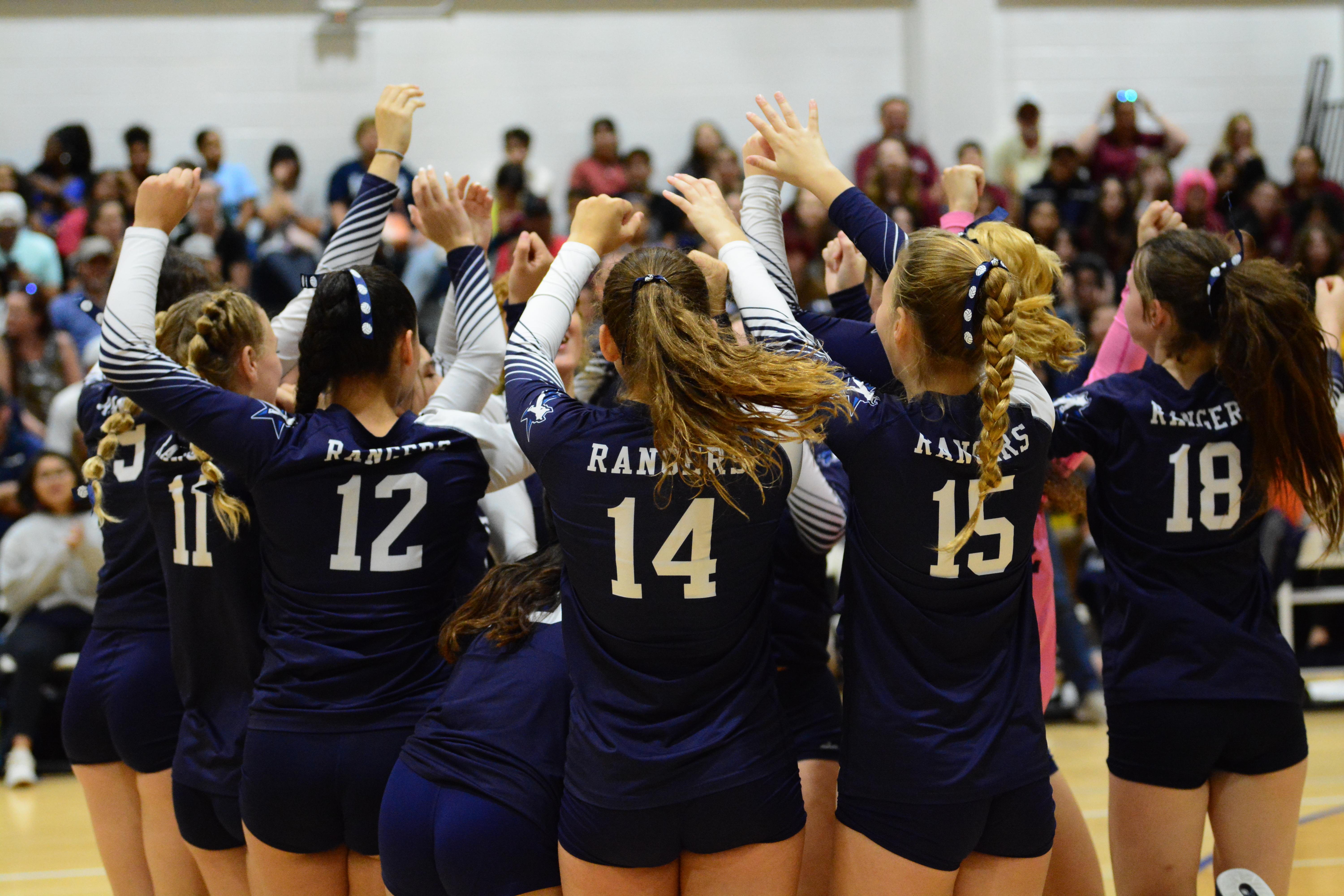 The volleyball athletes huddled together on the court, with their hands up in the air, wearing ribbons on their hair. From left to right: volleyball uniform numbers 9, 11, 12, 14, 15, and 18.