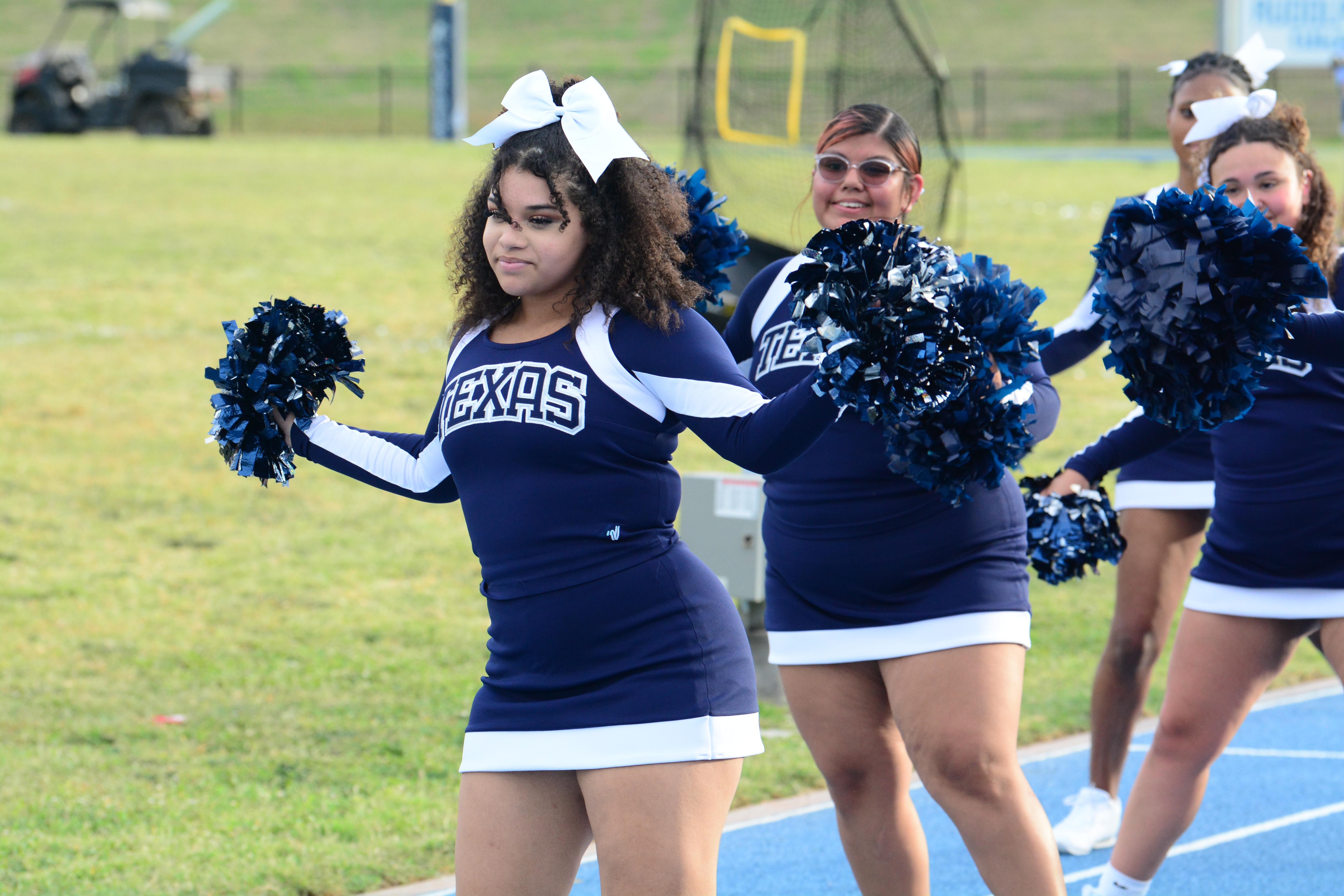 3 cheerleaders cheering for the football team, the girl in the front with black curls and white bow, wearing cheerleader uniform with “Texas” on the front.