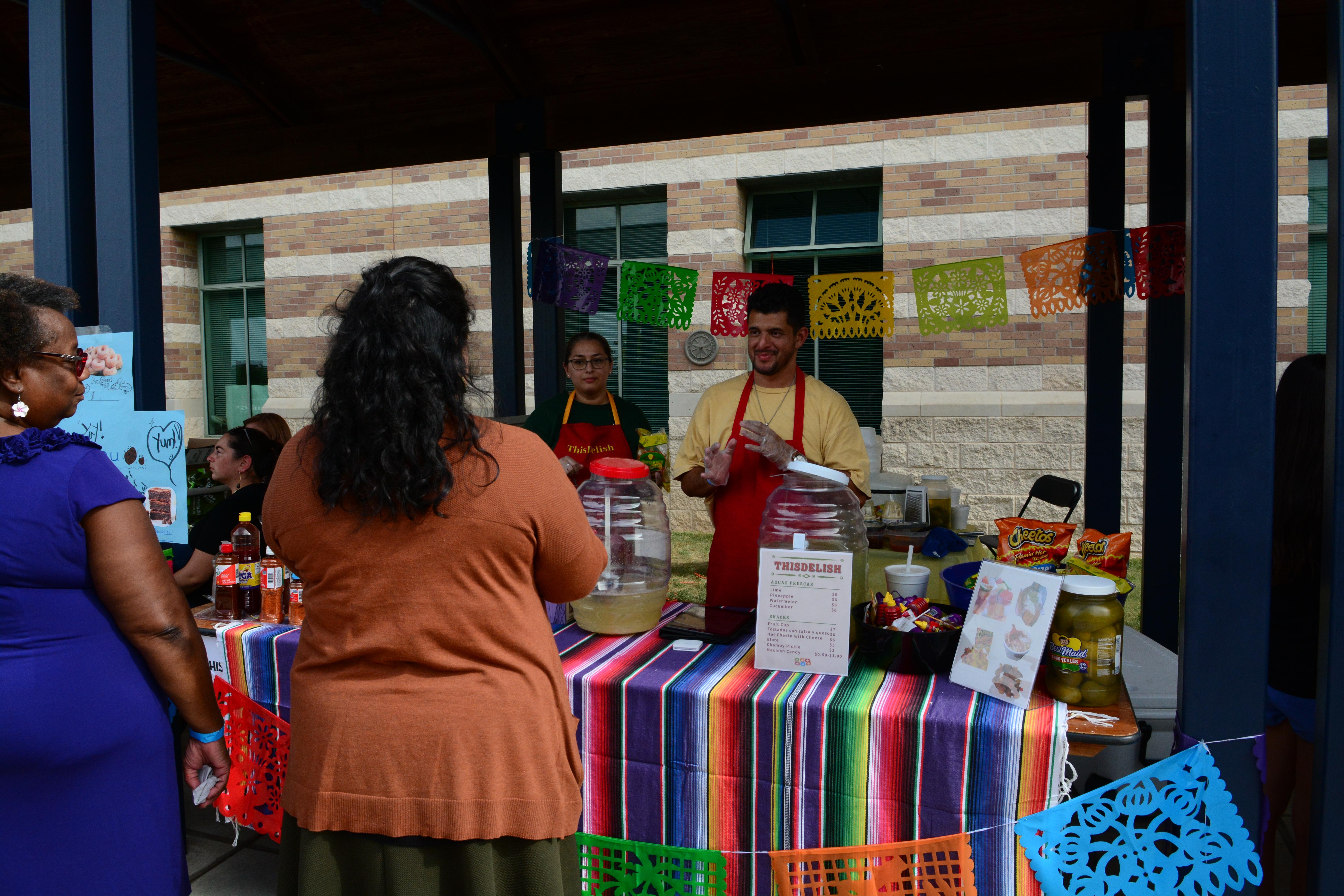 A Homecoming Booth, with a mexican blanket on the table, selling food, two people in red aprons talking with two customers.