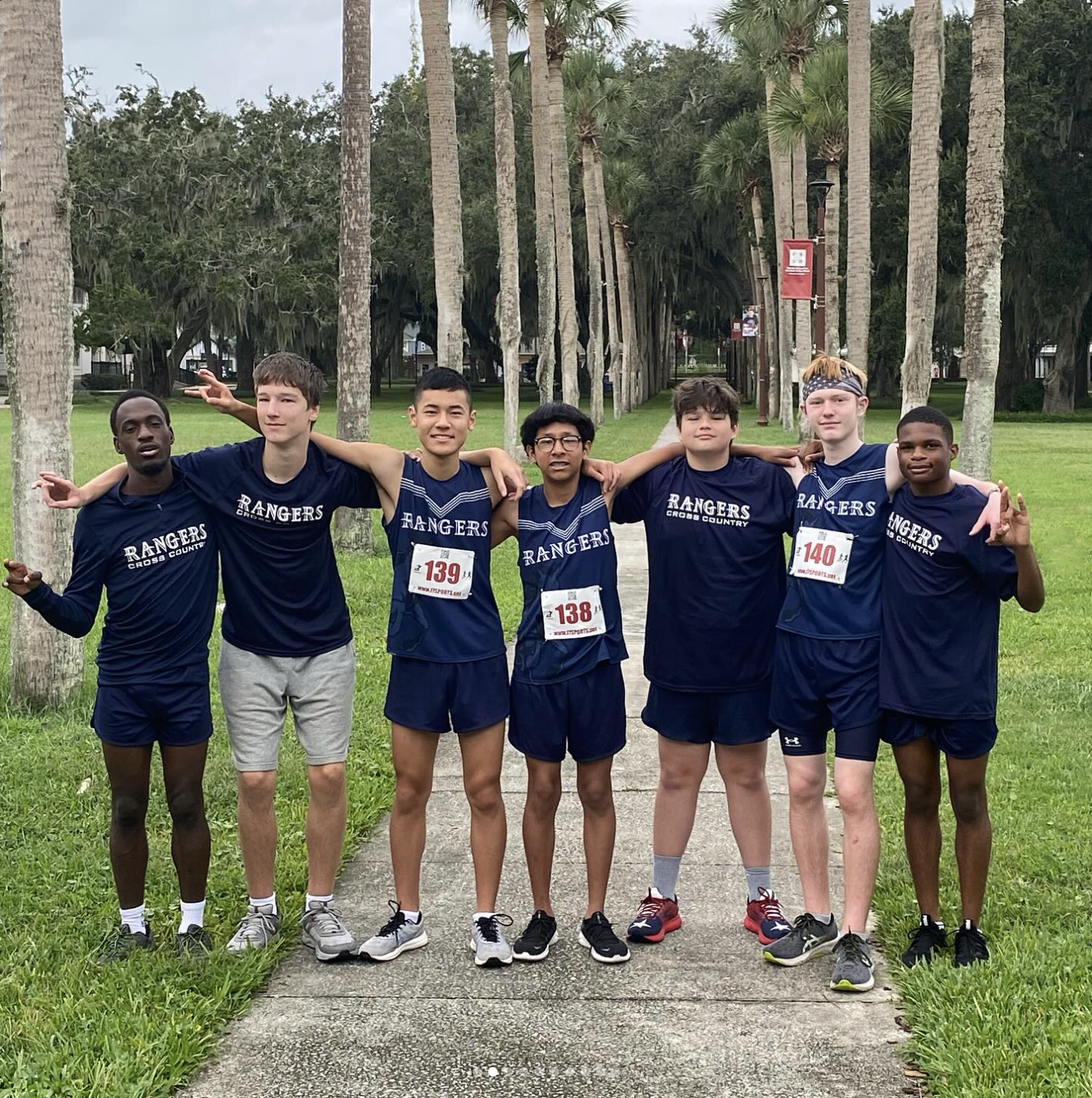 7 cross country athletes posed (some were smiling and some were not) behind two rows of palm trees and grass backgrounds. The athletics wore cross country numbered uniforms with words, “Rangers Cross Country” (navy blue and white).