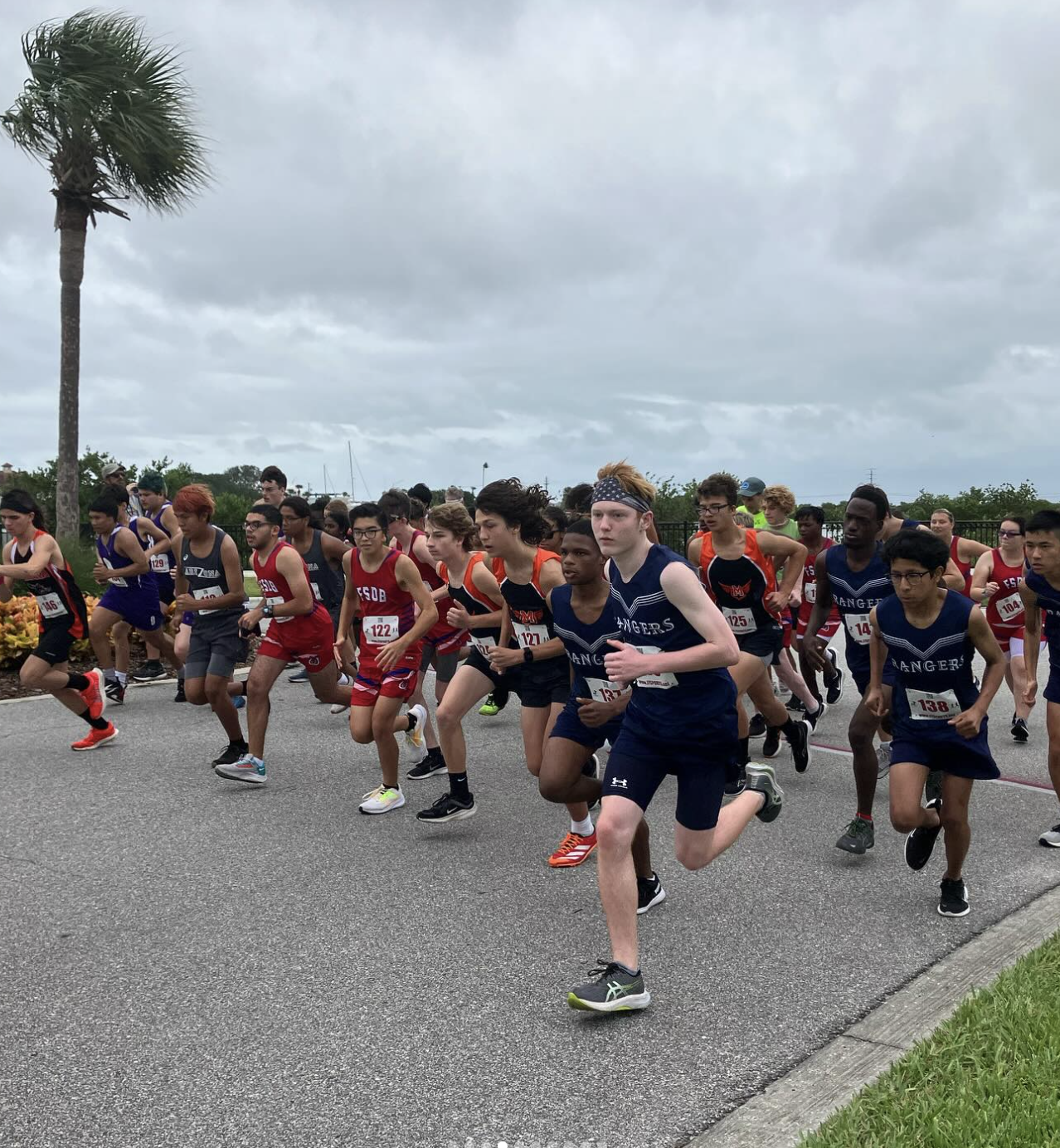 The background is cloudy with one palm tree. Different teams with different colors (red, navy, orange, and black) are on run. 