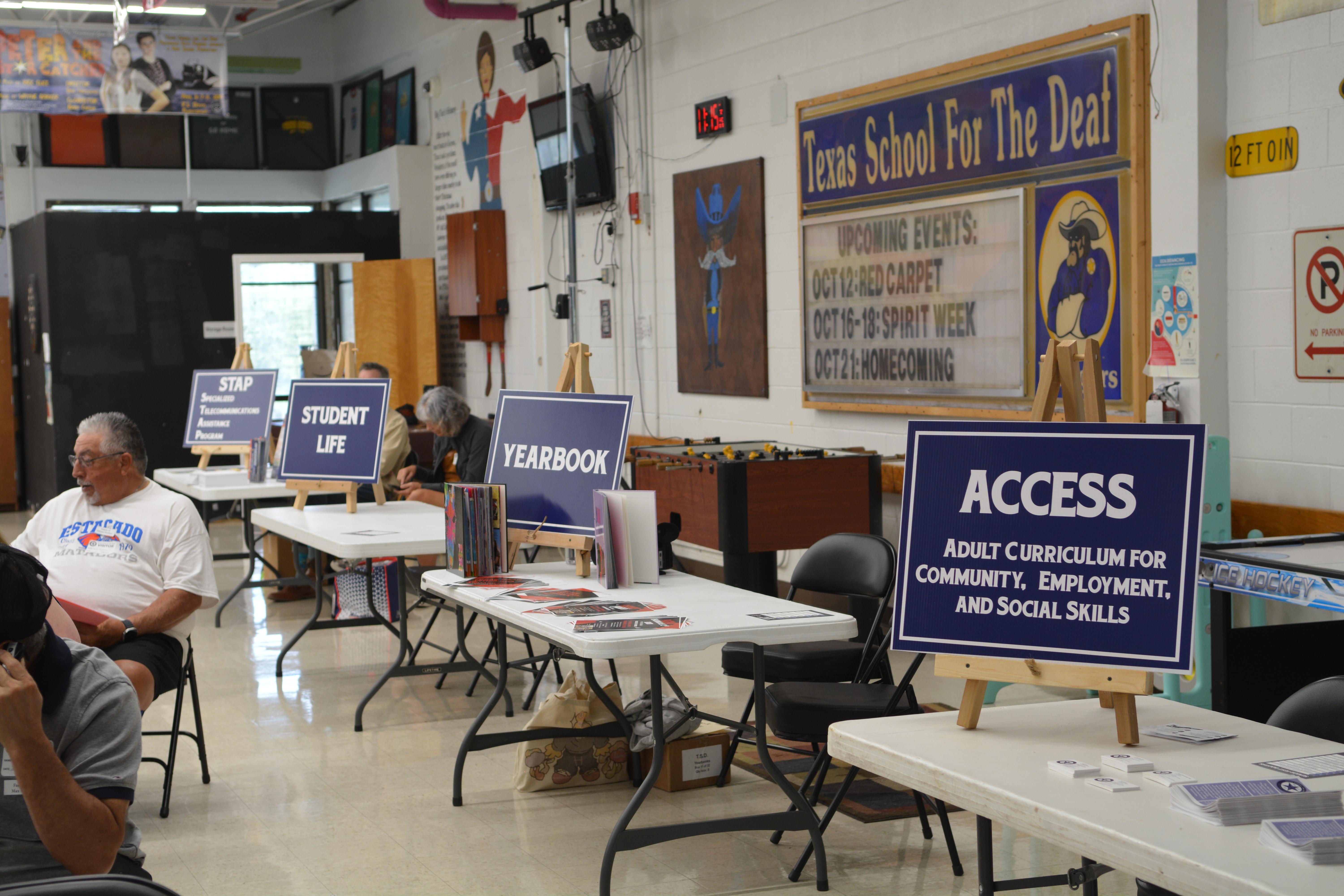 4 tables with poster stands in the order from front to back: ACCESS Adult Curriculum for Community, Employment, and Social Skills, Yearbook, Student Life, and lastly, STAP Specialized Telecommunications Assistance Program with people in the background.